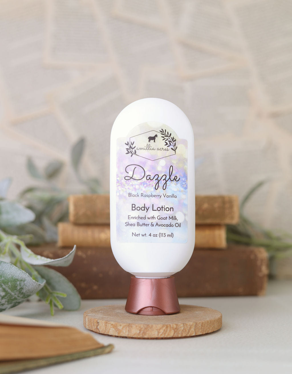 A bottle of Amillia Acres Dazzle Goat Milk Lotion sits on a round wooden stand, with blurred books and green leaves in the background. The label features shea butter, avocado oil, and highlights this rich handmade moisturizer.