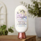 A bottle of Dazzle Goat Milk Lotion by Amillia Acres ~ Goat Milk Soaps and Lotions, featuring a floral label, sits atop stacked books with blurred green plants in the background.