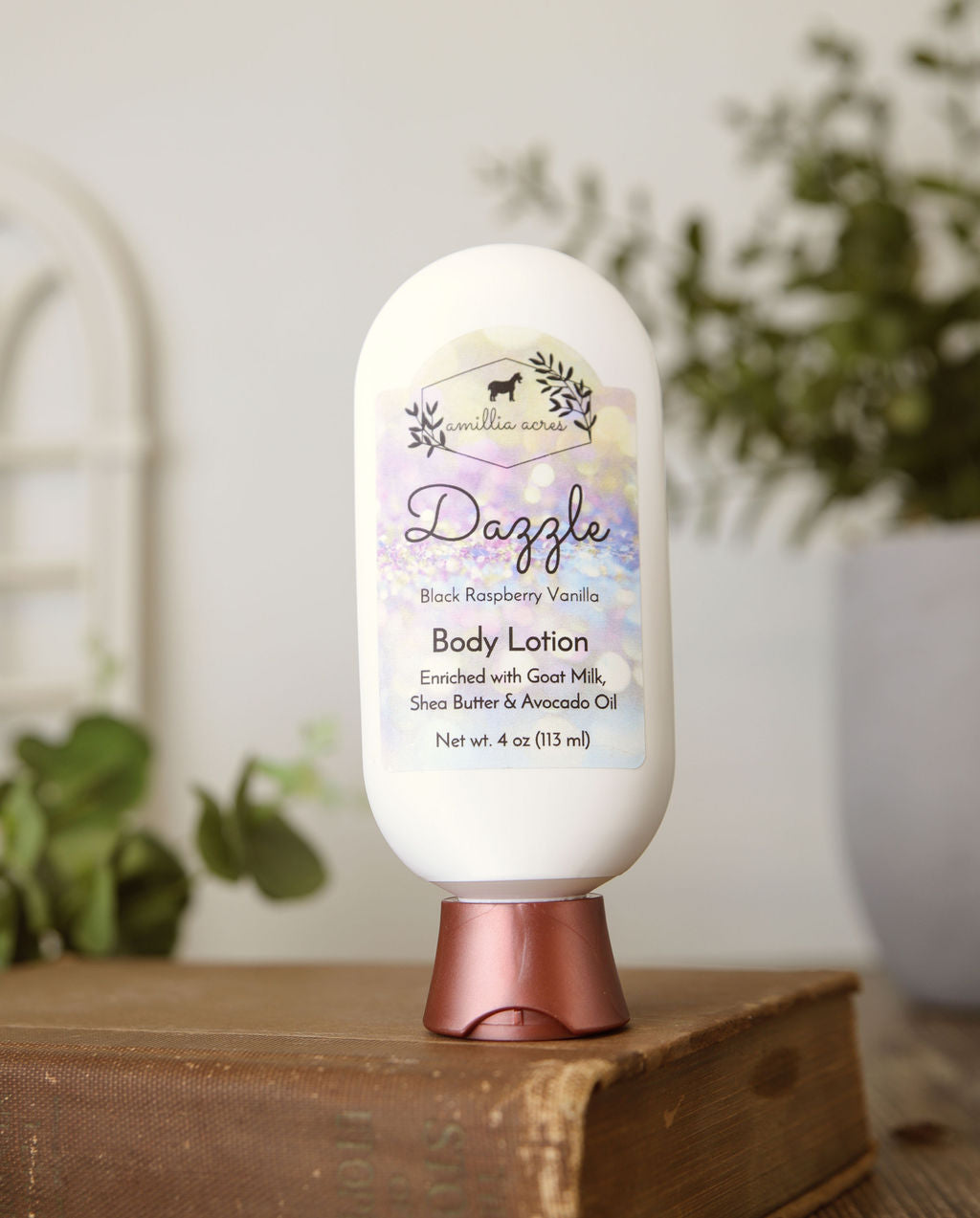 A bottle of Dazzle Goat Milk Lotion by Amillia Acres ~ Goat Milk Soaps and Lotions, featuring a floral label, sits atop stacked books with blurred green plants in the background.