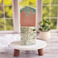 Two Escape artisanal soaps by Amillia Acres, infused with a delightful fragrance, feature swirled patterns and glitter on a white stand with wooden legs. A vibrant window offers a green view, accompanied by a small potted plant—ideal for handmade soap enthusiasts.