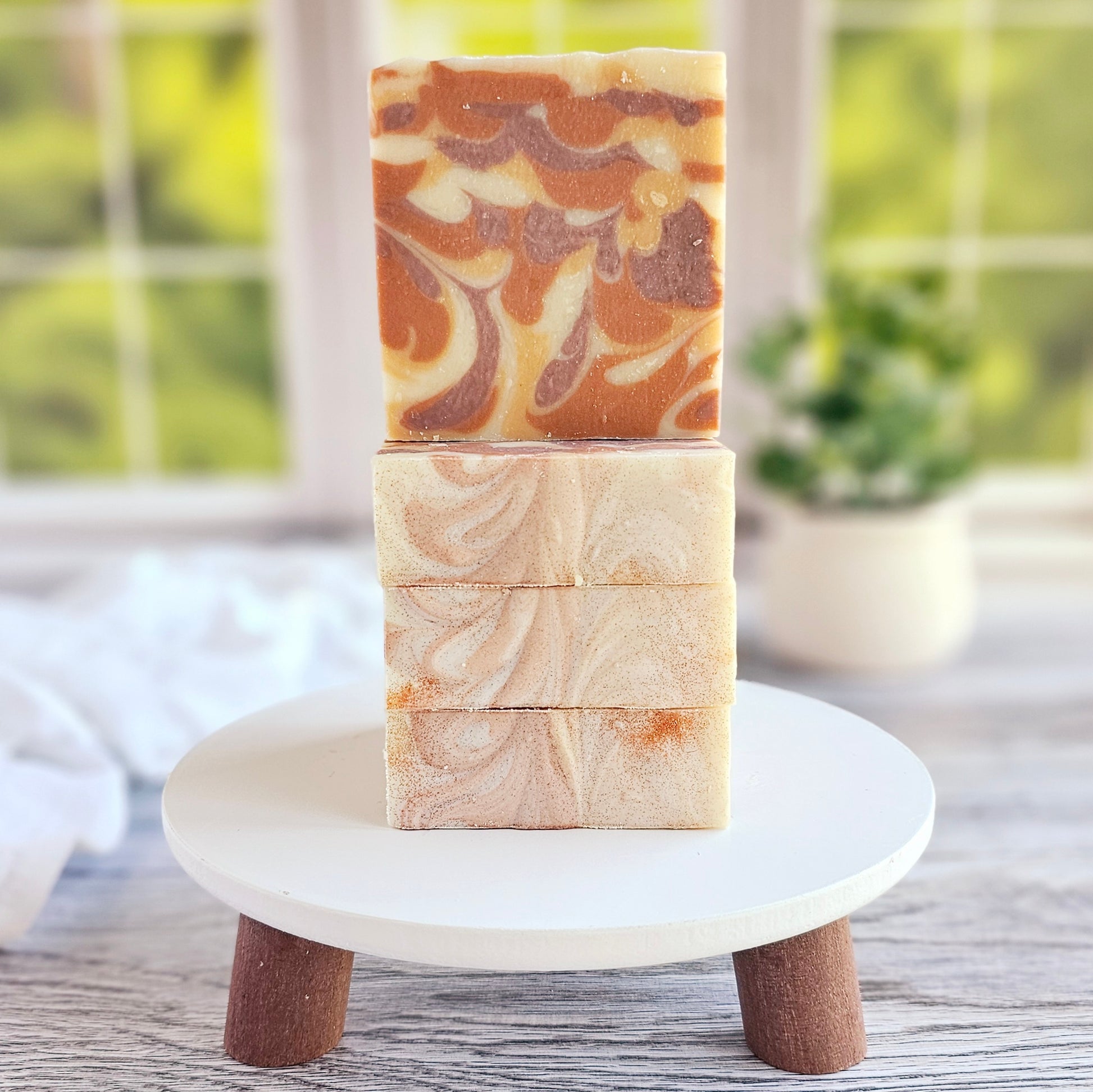 Three Amillia Acres Comfort Goat Milk soaps, made with natural oils, are stacked on a small white stand with wooden legs. The top bar showcases a swirled brown and cream design. Blurred green foliage and a window appear in the background.