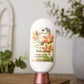 A bottle of "Happiness" Goat Milk Lotion by Amillia Acres ~ Goat Milk Soaps and Lotions, topped with a floral label, rests on a wooden surface, the background softly blurred with green plants and a white decorative window.