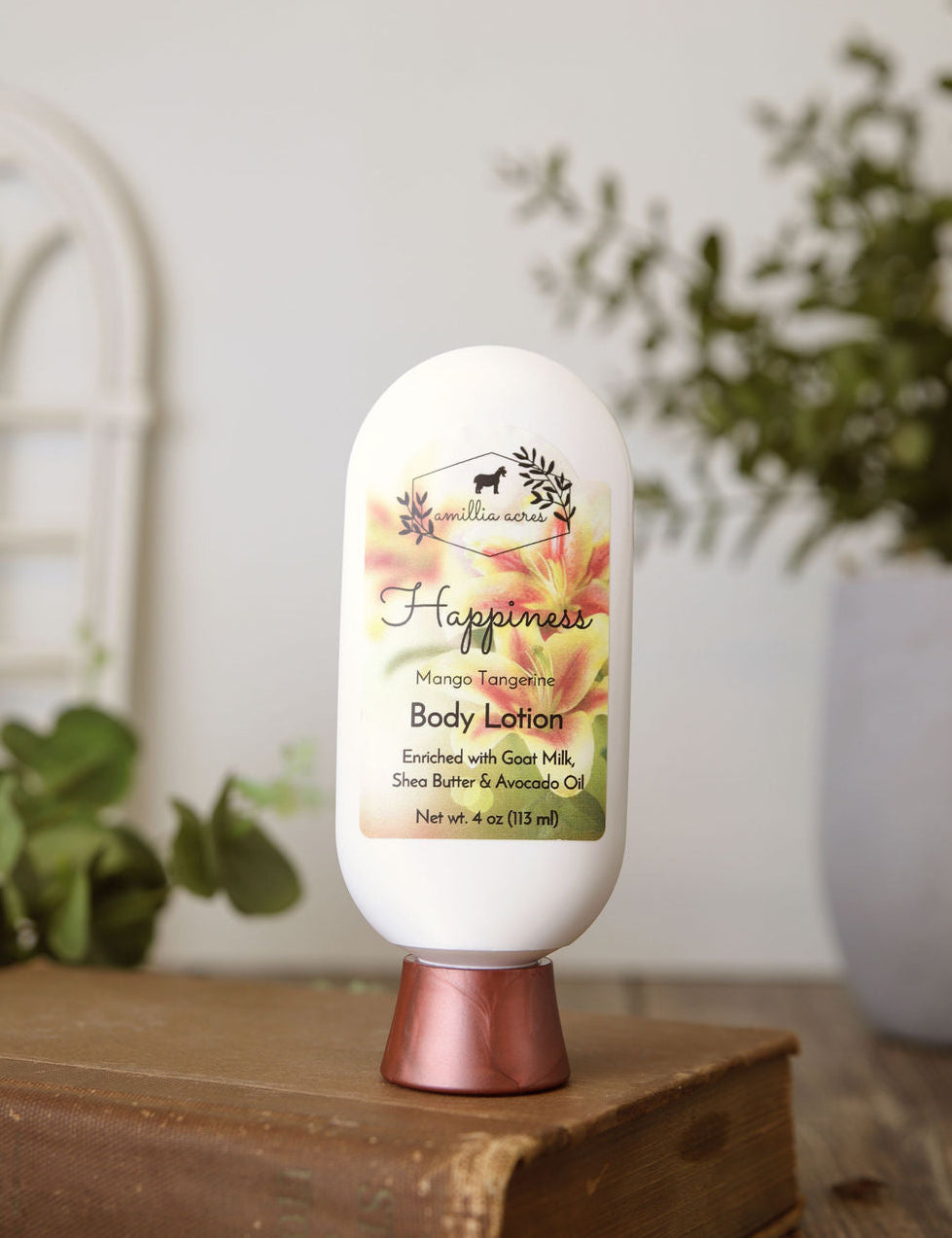 A bottle of "Happiness" Goat Milk Lotion by Amillia Acres ~ Goat Milk Soaps and Lotions, topped with a floral label, rests on a wooden surface, the background softly blurred with green plants and a white decorative window.