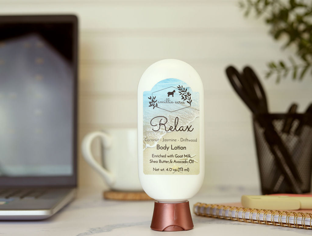 A bottle of Relax Goat Milk Lotion by Amillia Acres ~ Goat Milk Soaps and Lotions sits on a desk near a laptop, cup, notebook, and pens. Its label features coconut, jasmine, and driftwood scents for a luxurious moisturizing experience.