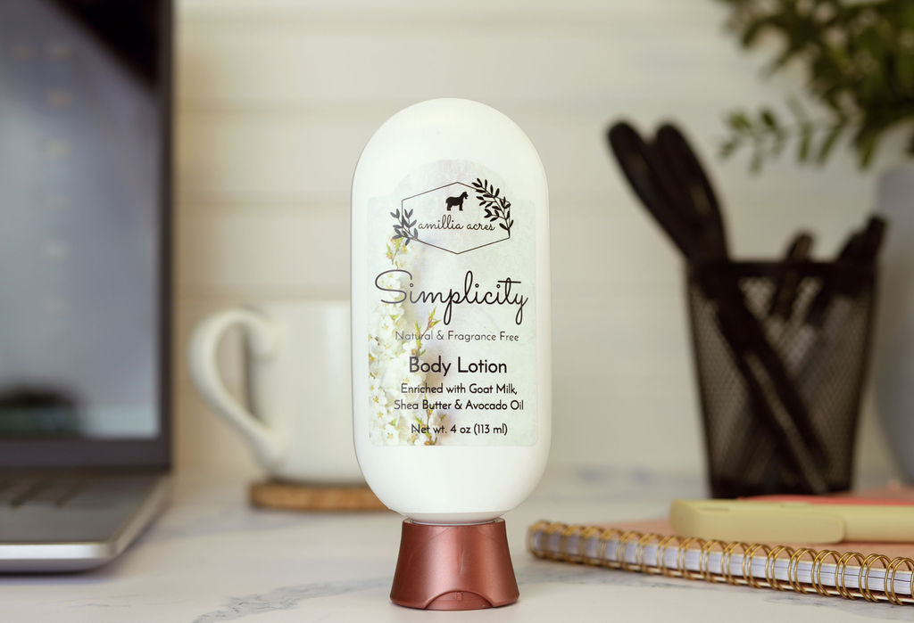 A bottle of Amillia Acres Simplicity Goat Milk Lotion sits on a desk beside a laptop, notebook, and cup of pens. Its label features nourishing goat milk, shea butter, and avocado oil, illuminated by gentle natural light.