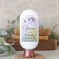 A white bottle of Amillia Acres Grace Goat Milk Lotion with a decorative label sits on a wooden stand, surrounded by greenery and stacked books in the background.