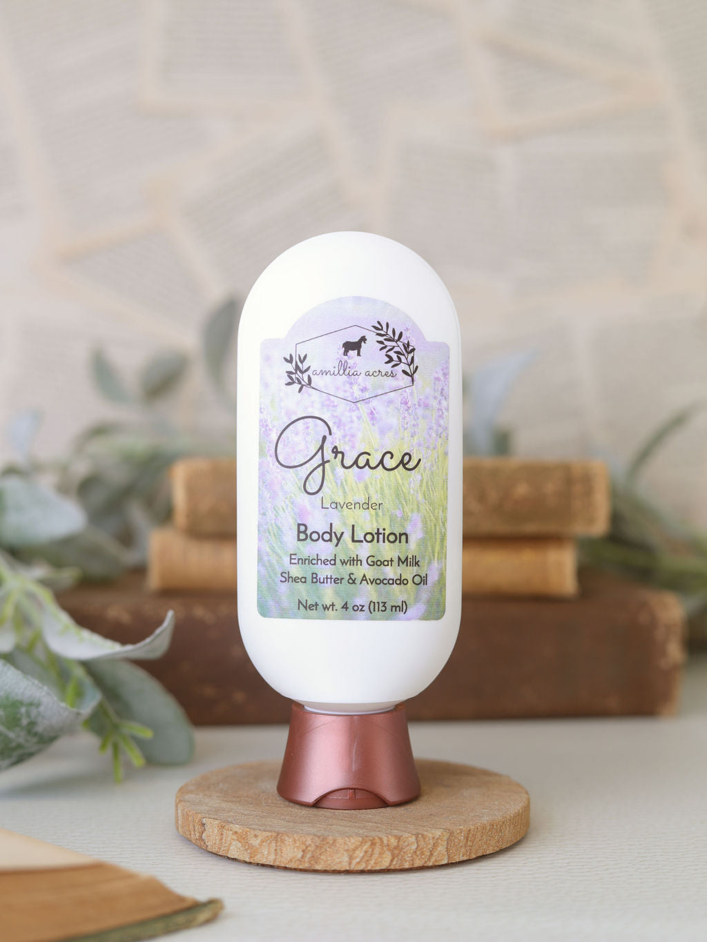A white bottle of Amillia Acres Grace Goat Milk Lotion with a decorative label sits on a wooden stand, surrounded by greenery and stacked books in the background.