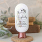 A white bottle of Amillia Acres Simplicity Goat Milk Lotion with a rose gold cap stands upright on a round wooden base, surrounded by stacked books and papers in a soft, natural setting.
