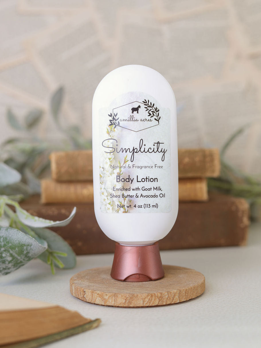 A white bottle of Amillia Acres Simplicity Goat Milk Lotion with a rose gold cap stands upright on a round wooden base, surrounded by stacked books and papers in a soft, natural setting.