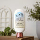 A bottle of Amillia Acres Relax Goat Milk Lotion sits on a closed book, surrounded by green plants and a white background. The label notes coconut, jasmine, driftwood, shea butter, and avocado oil for a luxurious moisturizing experience.
