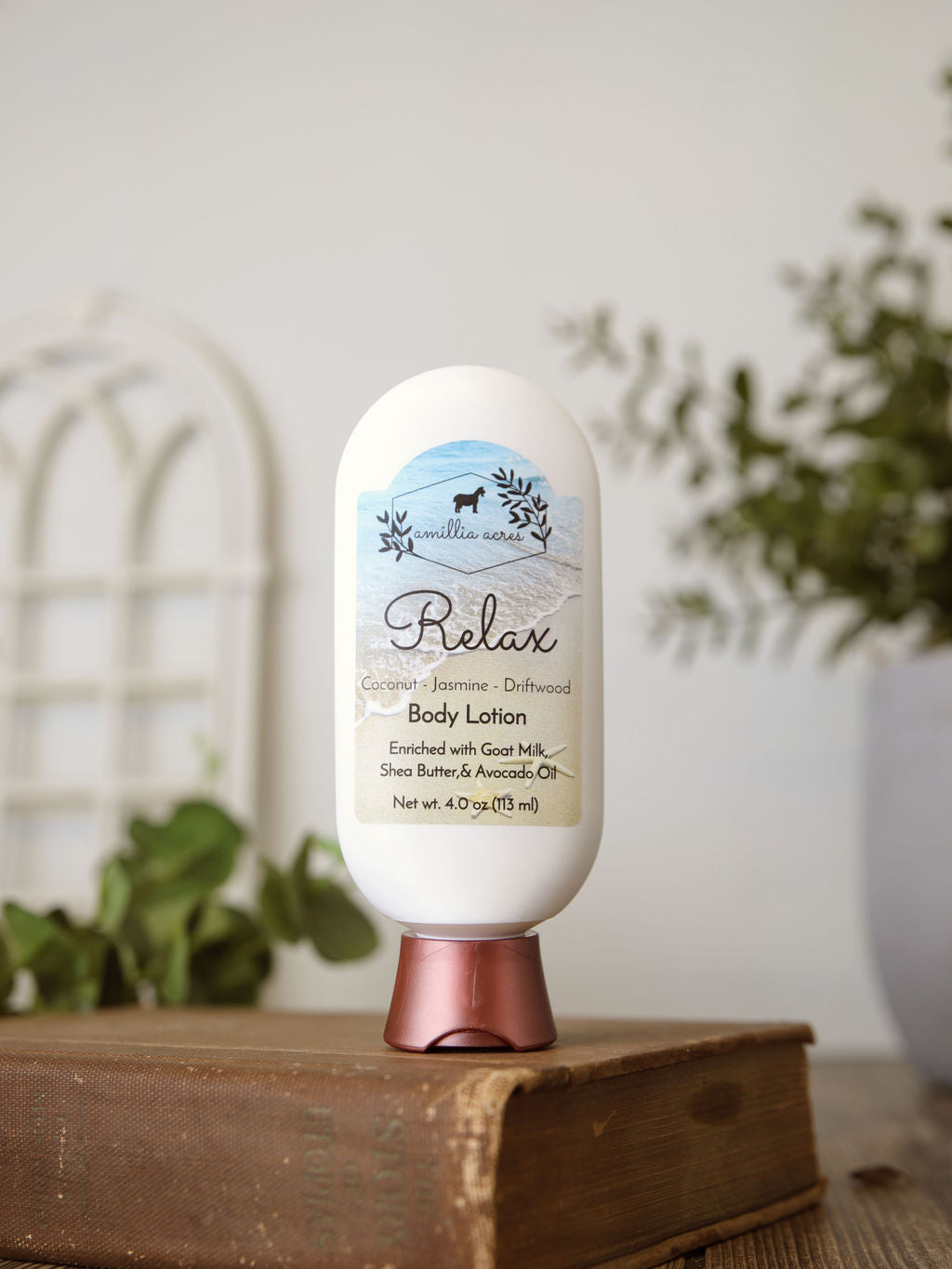 A bottle of Amillia Acres Relax Goat Milk Lotion sits on a closed book, surrounded by green plants and a white background. The label notes coconut, jasmine, driftwood, shea butter, and avocado oil for a luxurious moisturizing experience.