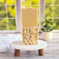 Three "I LOVE This Bar" goat milk soaps from Amillia Acres are stacked on a small white stand with wooden legs. A window showcasing blurry green foliage and a potted plant is in the background, enhancing the soap's natural, rustic texture and artisanal charm.