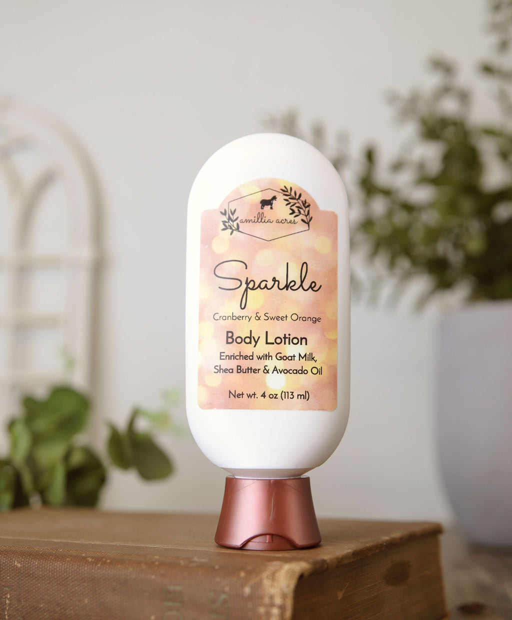 A bottle of Amillia Acres Sparkle Goat Milk Lotion sits on a wooden surface. The label reads "Cranberry & Sweet Orange" and features goat milk, shea butter, and avocado oil. A blurred plant appears in the background.