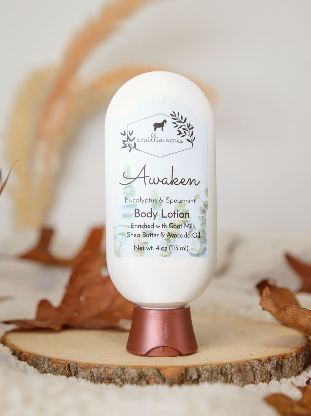 A white bottle of Amillia Acres Awaken Goat Milk Lotion with eucalyptus and spearmint rests on a wood slice among dried leaves, featuring goat milk, shea butter, and avocado oil against a neutral background.