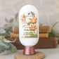 A bottle of Amillia Acres’ "Happiness" Goat Milk Lotion rests on a wooden stand, enriched with shea butter and avocado oil, surrounded by soft greenery with blurred books in the background.