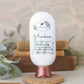 A white bottle of Amillia Acres Awaken Goat Milk Lotion with eucalyptus, spearmint, shea butter, and nyamplung oil stands on a wooden base. Books and green foliage form a softly blurred background.
