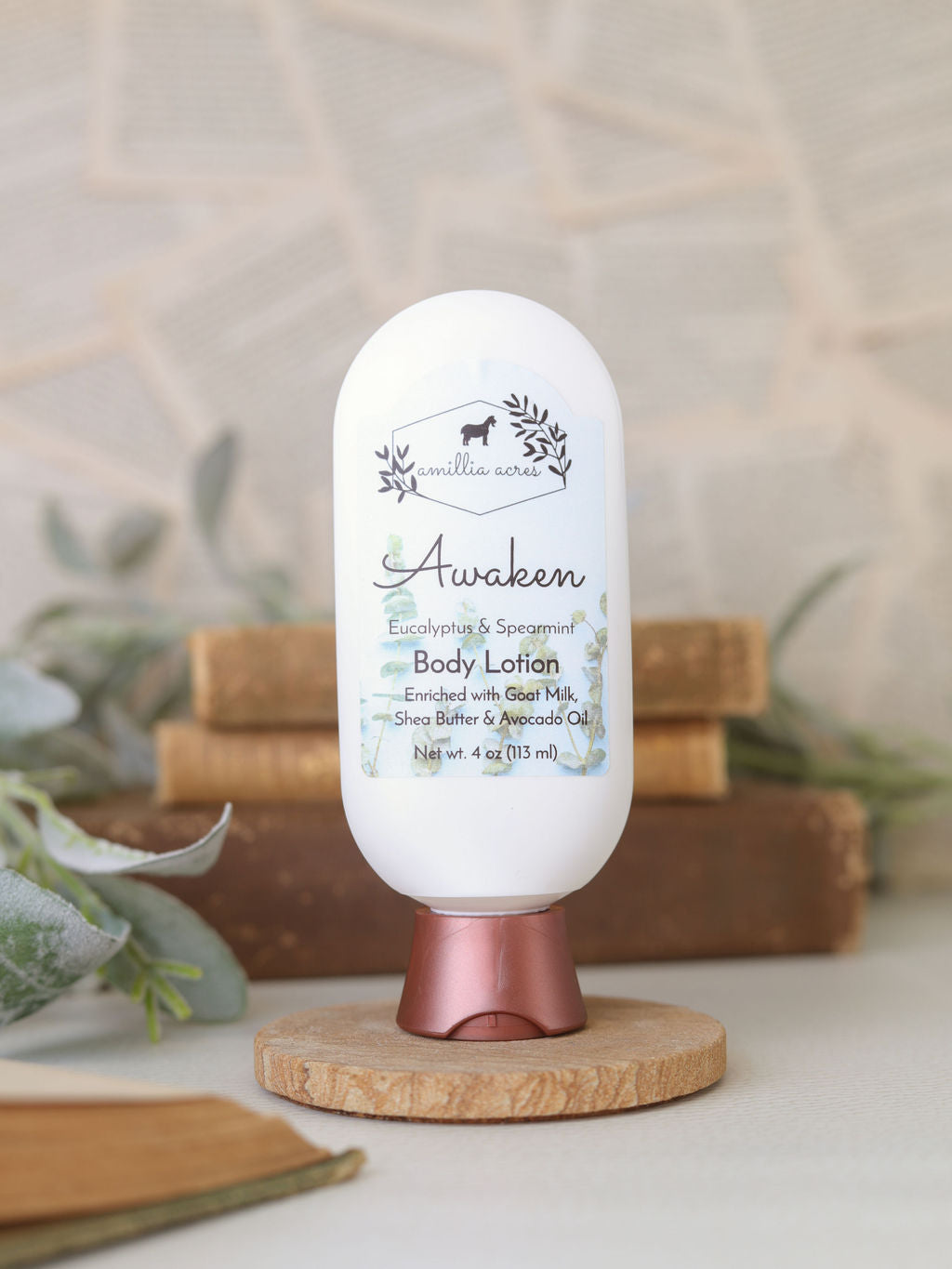 A white bottle of Amillia Acres Awaken Goat Milk Lotion with eucalyptus, spearmint, shea butter, and nyamplung oil stands on a wooden base. Books and green foliage form a softly blurred background.