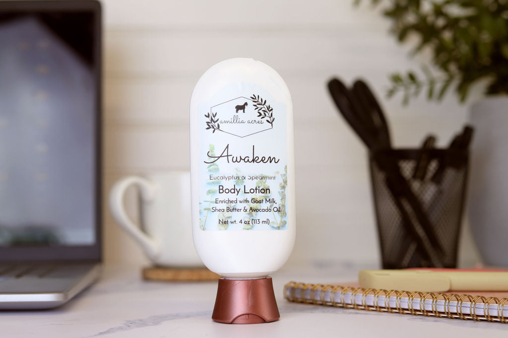 An Amillia Acres Awaken Goat Milk Lotion sits on a desk near a laptop and notebook, its label highlighting nourishing goat milk, shea butter, and avocado oil.
