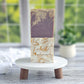 Grace Goat Milk Soap by Amillia Acres features artful swirls in purple, cream, and brown on a white stand with wooden legs. The blurred window background highlights the lavender-scented soap nestled next to a potted plant.