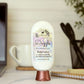 A bottle of Amillia Acres Dazzle Goat Milk Lotion sits on a desk next to a spiral notebook, cup, and pen container, with a laptop partly visible in the background.