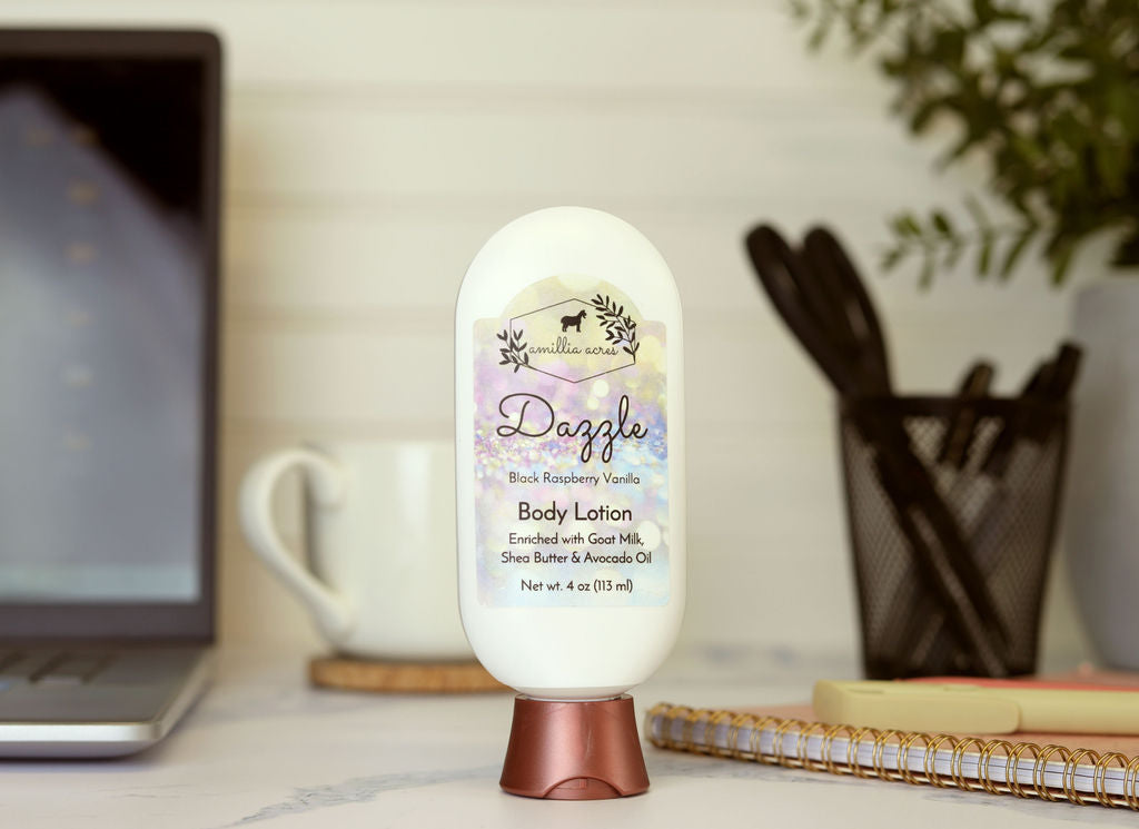 A bottle of Amillia Acres Dazzle Goat Milk Lotion sits on a desk next to a spiral notebook, cup, and pen container, with a laptop partly visible in the background.