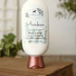 A white bottle of Amillia Acres Awaken Goat Milk Lotion, with eucalyptus and spearmint plus shea butter and avocado oil, stands on a closed book against a blurred background.