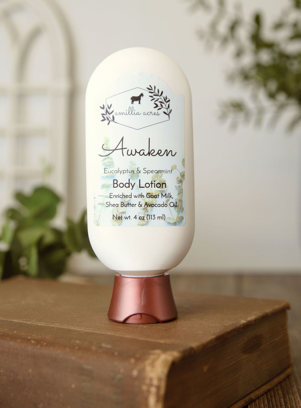 A white bottle of Amillia Acres Awaken Goat Milk Lotion, with eucalyptus and spearmint plus shea butter and avocado oil, stands on a closed book against a blurred background.