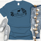 The Amillia Acres ~ Goat Milk Soaps and Lotions short sleeve t-shirt, made from 100% preshrunk cotton, features a charming goat and floral design. It effortlessly complements gray sneakers, faded blue jeans, and a natural touch as it sits gracefully on a white wooden background.