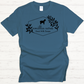 The Amillia Acres ~ Goat Milk Soaps and Lotions T-shirt, crafted from 100% preshrunk cotton, showcases a Gildan Softstyle fit in blue with a black goat silhouette set among leaves. It features the text "Amillia Acres Goat Milk Soaps" within a hexagonal frame and is displayed on a light wooden surface, combining comfort with rustic charm.