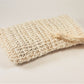 On a white surface rests the Exfoliating Soap Saver Bag by Amillia Acres, a textured beige scrubber crafted from natural sisal with a convenient looped handle. Ideal for gentle exfoliation, it also serves as an exfoliating soap saver.