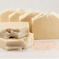 Five bars of Amillia Acres Simplicity Goat Milk Soap are artfully arranged on a white surface: two bars stacked, one standing, and two leaning. Pink salt grains scattered nearby add texture and enhance the soap's moisturizing qualities in this serene composition.