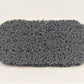 The Amillia Acres Soap Saver is a rectangular gray pad with a textured, curly surface that looks like tightly wound loops of wire, set against a plain white background to help maximize the lifespan of your soap.