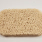 Amillia Acres' Soap Saver is a rectangular beige loofah pad featuring a rough, intertwined texture on a white background, designed to extend the life of your soap.