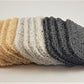 A collection of Amillia Acres Soap Saver pads is displayed in four elegant color variations: beige, light gray, white, and dark gray. Each stack features multiple pads arranged in an overlapping manner against a neutral backdrop. These soap savers are crafted to enhance the longevity of your soap while adding a touch of style and functionality.