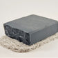 A dark gray, rectangular bar of soap with a rough, textured surface on one side is placed on the Amillia Acres "Soap Saver," a light, woven mat, set against a plain white background.