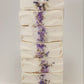 Grace by Amillia Acres is a handmade goat milk soap, cream-colored with a central stripe of dried lavender flowers and herbs. Its smooth texture features slight ridges, accentuated by natural oils for an artisanal look.