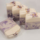 Five Grace goat milk soaps by Amillia Acres, in cream and purple swirls, are displayed. Four stand upright displaying a textured top with bits sprinkled on it, while one lies flat. These handmade lavender soaps have an appealing marbled pattern against a white background.