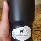 A hand holds a black insulated tumbler featuring an "Amillia Acres ~ Goat Milk Soaps and Lotions" vinyl sticker with a horse silhouette. The durable label contrasts against the speckled countertop, highlighted by the tiled backsplash.