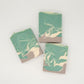 Three rectangular bars of Amillia Acres' "Relax | Goat Milk Soap" are artfully arranged on a light surface; two parallel and one diagonally between, showcasing a swirled green, white, and brown design infused with natural oils.