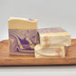 Three Grace | Goat Milk Soap bars from Amillia Acres, with purple and cream swirls, sit on a wooden tray. Two are stacked, one upright, each adorned with small purple and gold crystals, highlighting the luxurious essence of lavender goat milk soap.