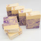 Six Grace goat milk soaps by Amillia Acres, featuring a cream and purple marbled design, are arranged in a staggered format on white. They are infused with natural oils, and their top edges have a dusting of purple and gold pigments, hinting at the lavender scent within.