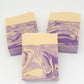 Three handmade rectangular soaps from Amillia Acres called "Grace" feature a swirled purple and cream design. Each lavender goat milk bar has a solid cream top, contrasting with the marbled pattern below, and is infused with natural oils resting elegantly on a plain white surface.