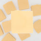 A close-up of a square piece of I LOVE This Bar | Goat Milk Soap by Amillia Acres ~ Goat Milk Soaps and Lotions, centered against a blurred background of soap bars arranged in a circular pattern. The light yellow soap resembles the richness and artisanal allure one might find in fine cheese.