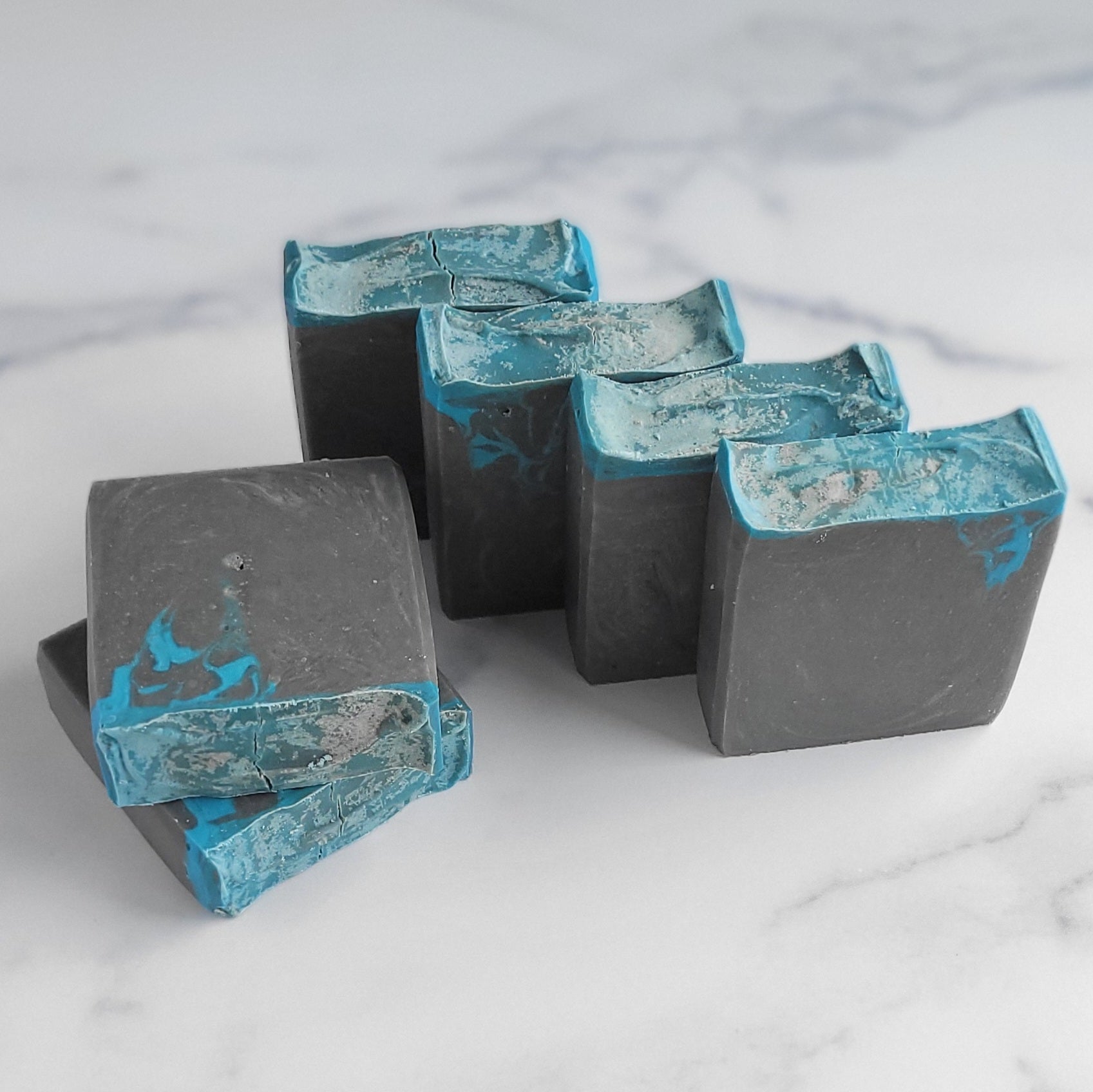 Five rectangular bars of Bay Rum Goat Milk Soap from Amillia Acres ~ Goat Milk Soaps and Lotions, featuring a marbled design in shades of black, gray, and blue, are neatly arranged on a light marble surface.