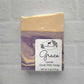 A square bar of Amillia Acres' Grace Goat Milk Soap, featuring purple and cream swirls, has a label with a goat and leaves stating "Grace Lavender Goat Milk Soap, Net wt. 4 oz (113 g)." Infused with nourishing natural oils, it is displayed against a white brick wall backdrop.