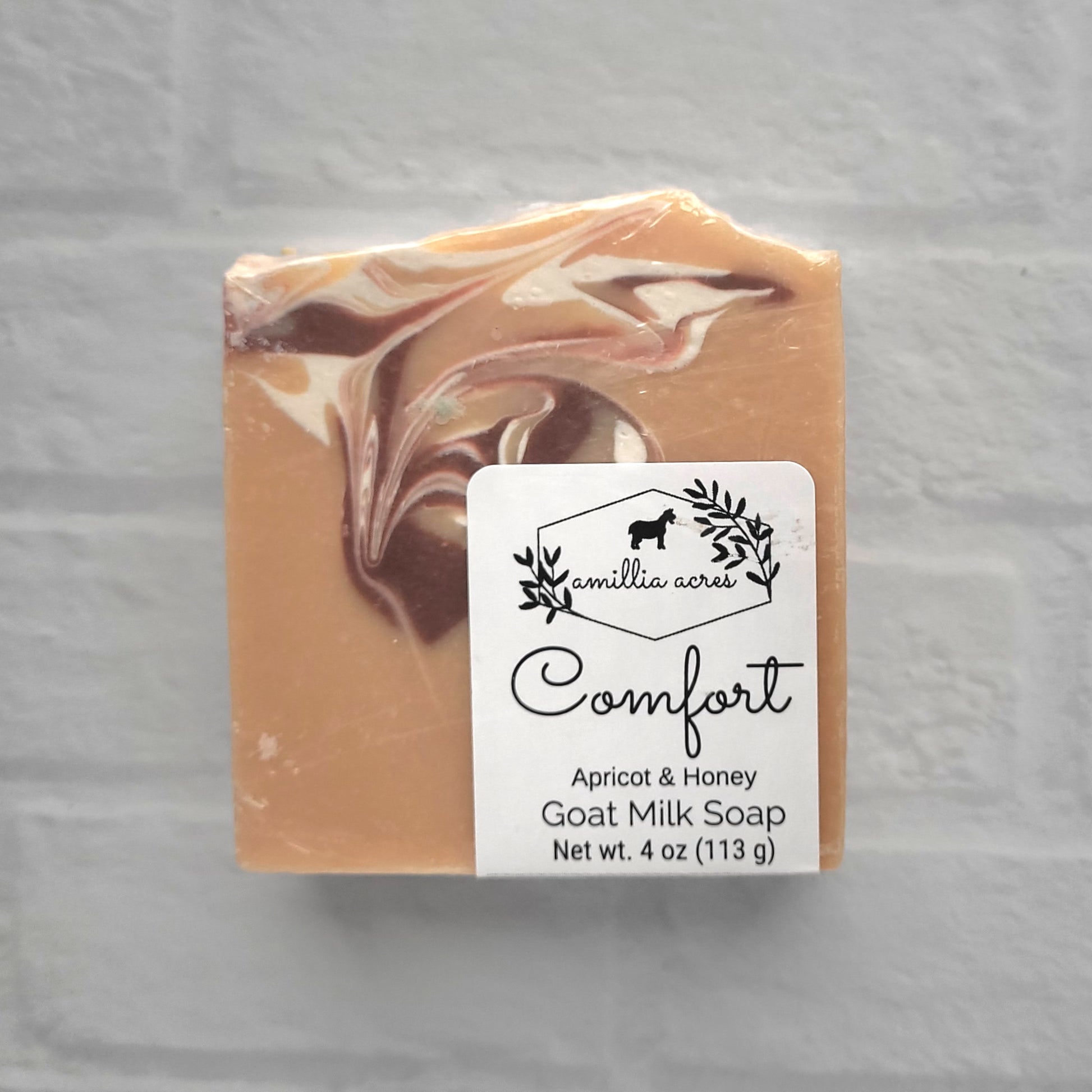 The "Comfort" goat milk soap by Amillia Acres is a 4 oz bar with an apricot and honey scent. It features simple packaging with a logo of leaves and a small animal, enhanced with natural oils against a light gray brick background.