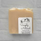 A beige, fragrance-free handmade goat milk soap bar, labeled "Amillia Acres Simplicity," weighing 4 oz (127 g), rests against a light gray brick background.