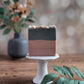 A bar of Sweater Weather Goat Milk Soap from Amillia Acres, featuring gray and peach layers, sits on a white pedestal. A blurred gray vase with flowers is in the background, while green leaves in the foreground harmonize with the natural oils infused within.