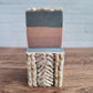 The Sweater Weather goat milk soap from Amillia Acres features intricate gray and pink swirls and braids, displayed atop another soap made from natural oils. The warm wooden surface contrasts against a blurred white brick wall backdrop.