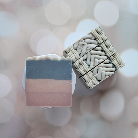 Amillia Acres' Sweater Weather Goat Milk Soaps: one with natural oils in gray, pink, and cream layers; the other with intricate swirls. A soft bokeh background accentuates these exquisite handmade soaps.